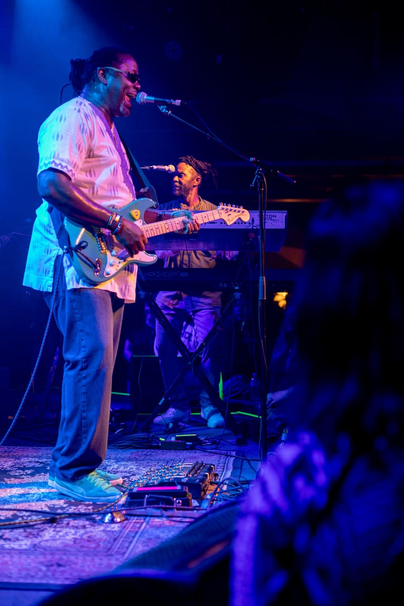 Musician singing and playing guitar on stage under blue and pink stage lighting with a keyboardist seated behind.