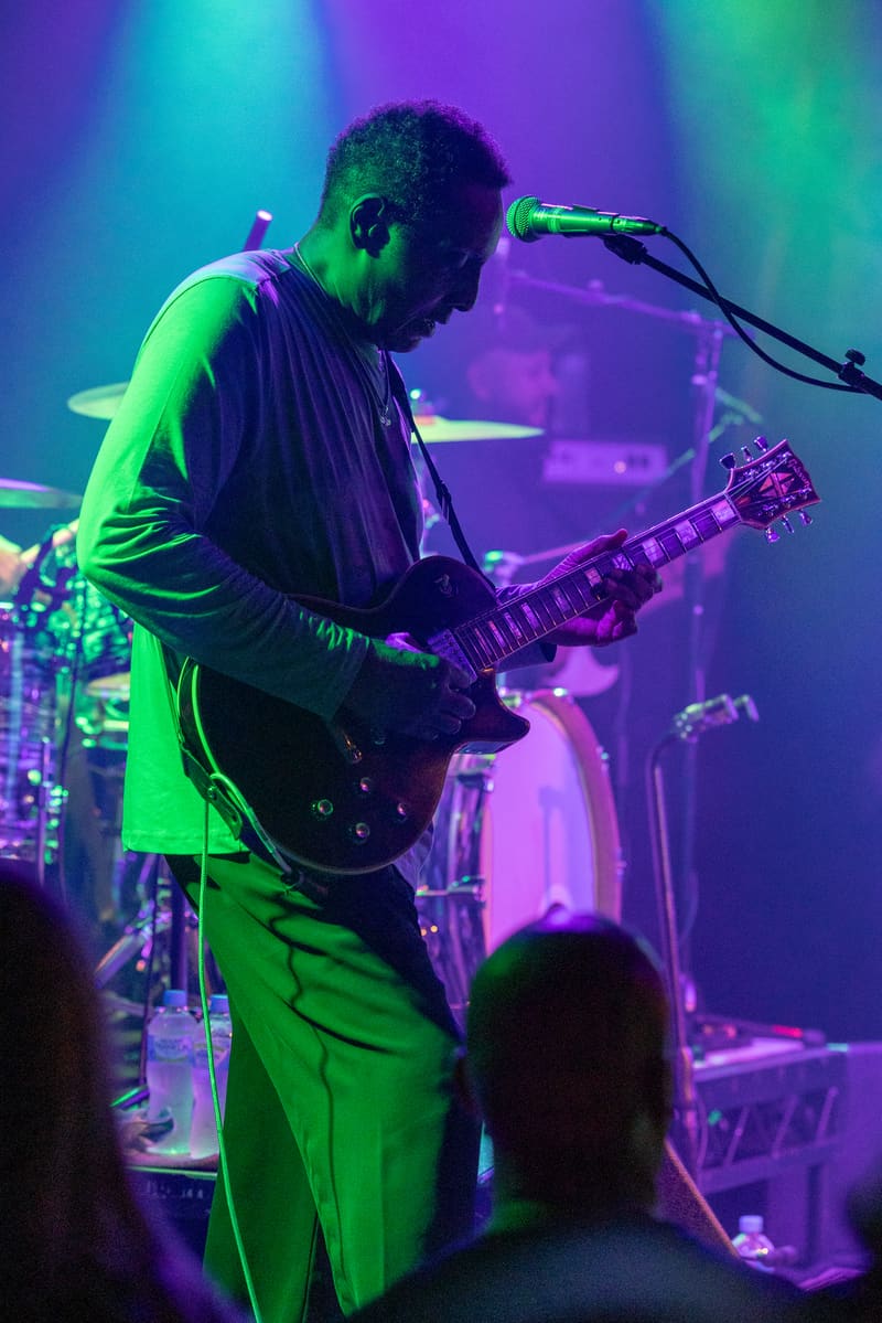 Guitarist performs at microphone under vibrant purple and green stage lighting during The Original Wailers concert.