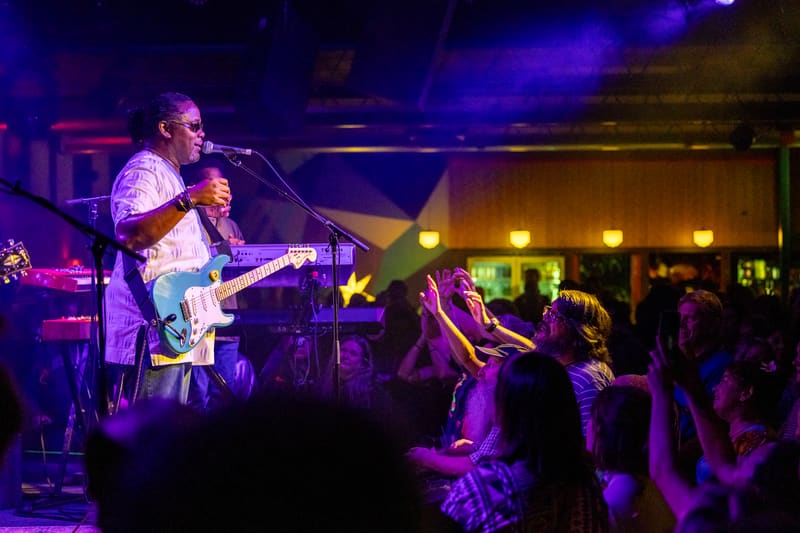 Guitarist and singer performing on stage under purple and yellow lights with audience members dancing in the foreground.