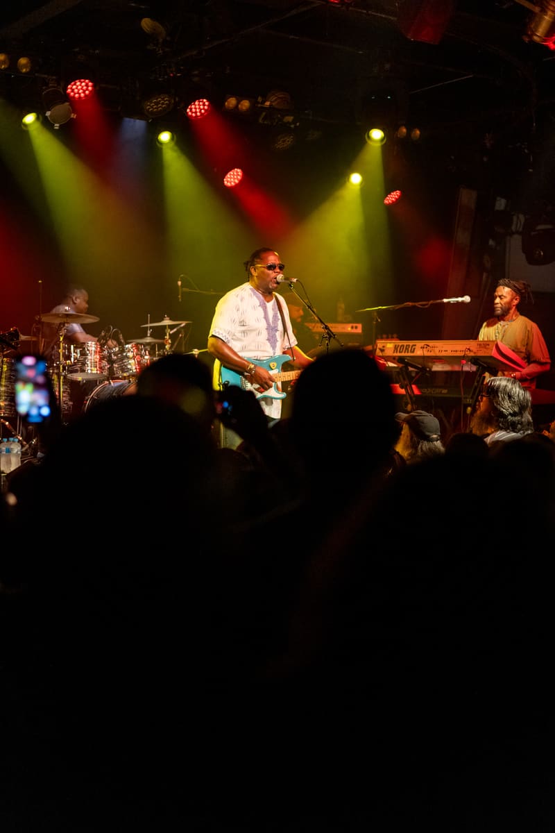 Guitarist in white shirt performing centre stage under red and yellow spotlights with band members and audience silhouettes visible.