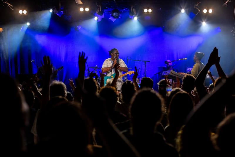 Performer on stage under blue spotlights with crowd of raised hands during live concert.