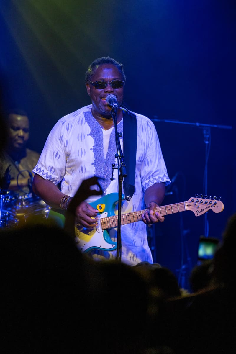 Guitarist and vocalist performs at the microphone with band members visible behind, stage lit in blue tones.
