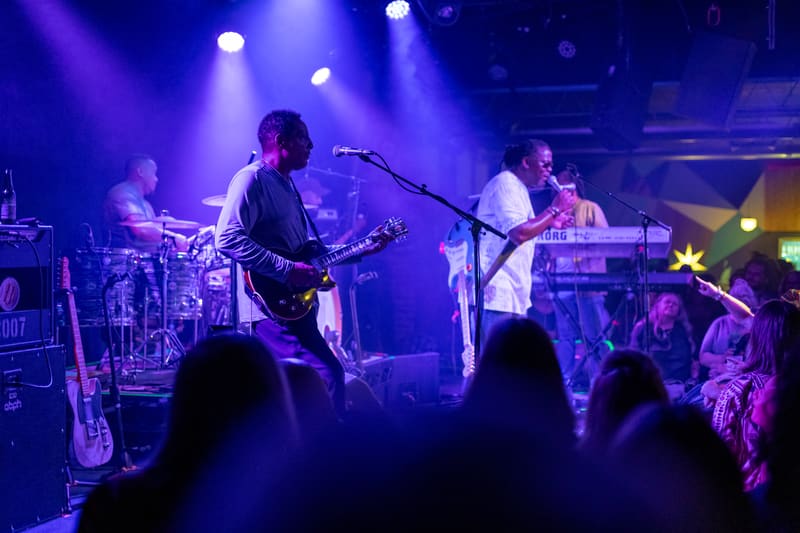 Musicians performing on stage with guitars and drums, bathed in blue and purple stage lighting, audience silhouettes in foreground.