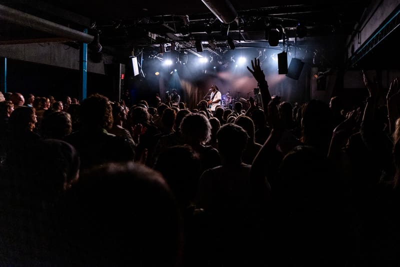 Packed crowd of concert-goers with hands raised beneath stage lights at a live music performance.