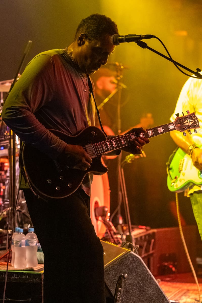 Guitarist singing into microphone at stage, bathed in warm yellow and orange stage lighting during a live performance.