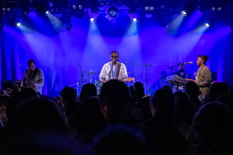 Band members perform on stage bathed in blue spotlights, audience silhouettes in foreground.