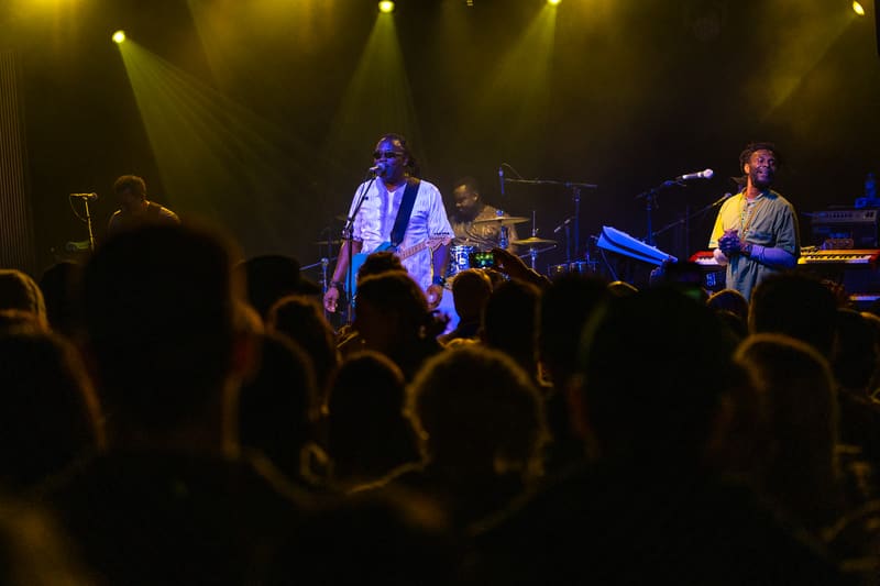 The Original Wailers perform on stage beneath warm spotlights, with audience members silhouetted in the foreground.