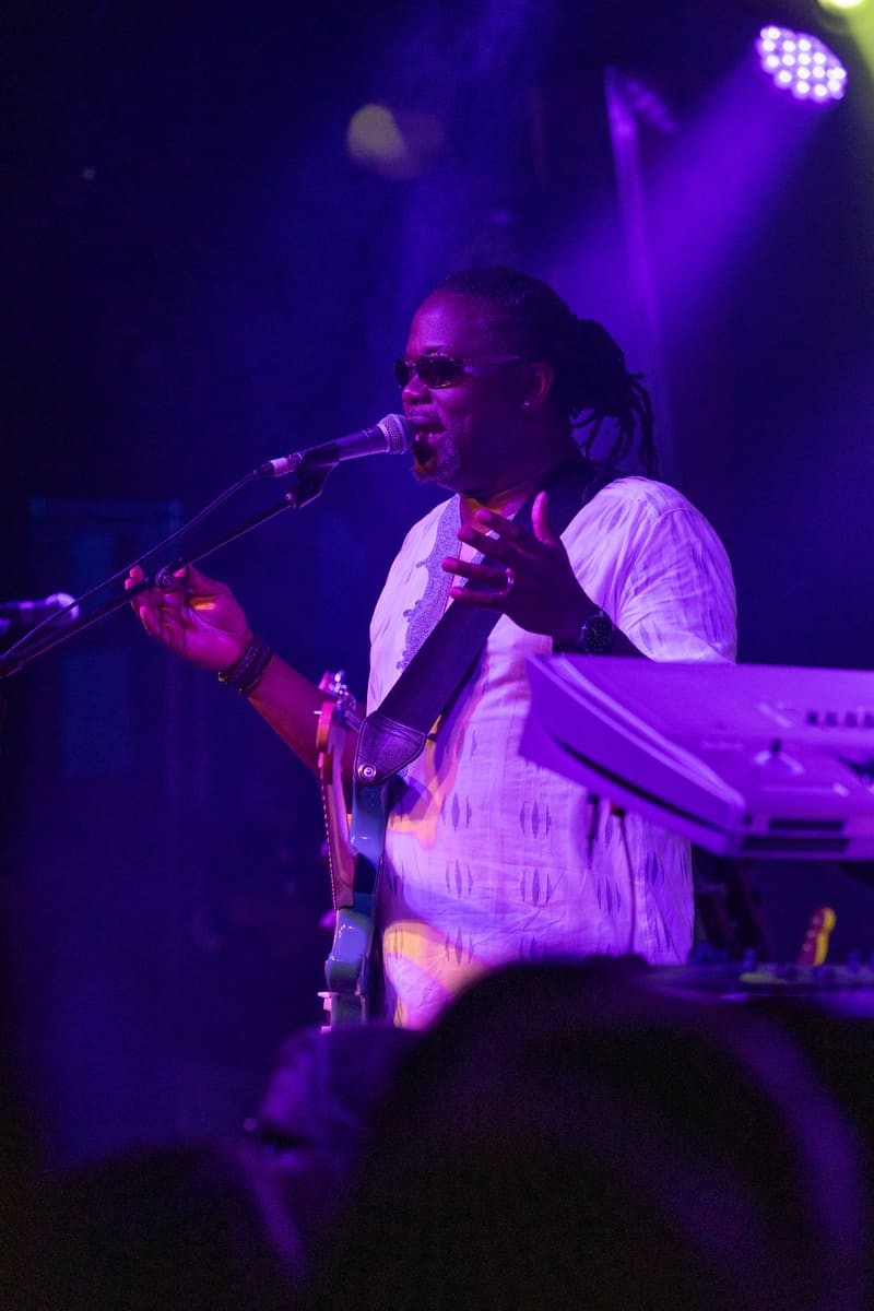 Musician performing at a microphone under purple stage lighting, wearing a white shirt and sunglasses.