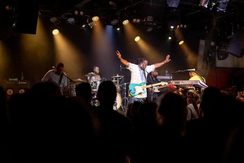The Original Wailers perform on stage with arms raised, bathed in warm spotlights before an engaged crowd.