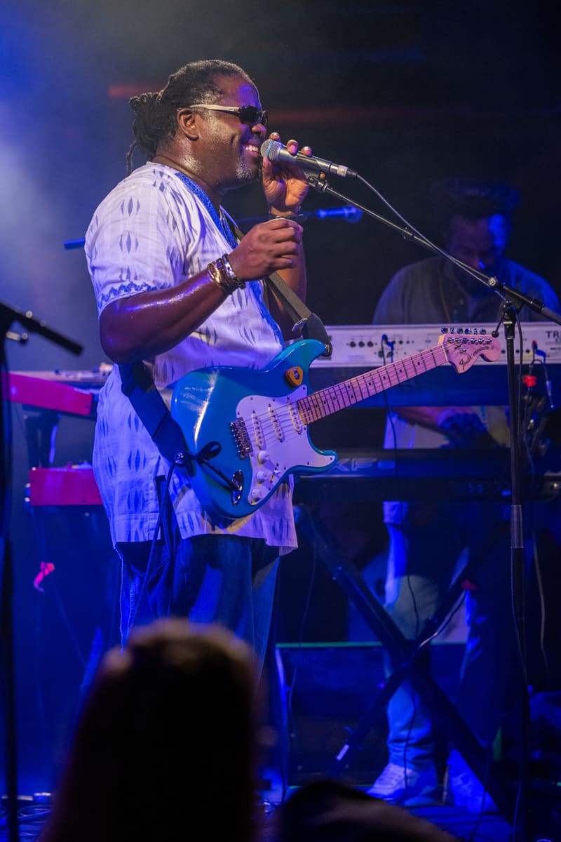 Musician performing at a microphone with bass guitar under blue stage lighting during The Original Wailers concert.