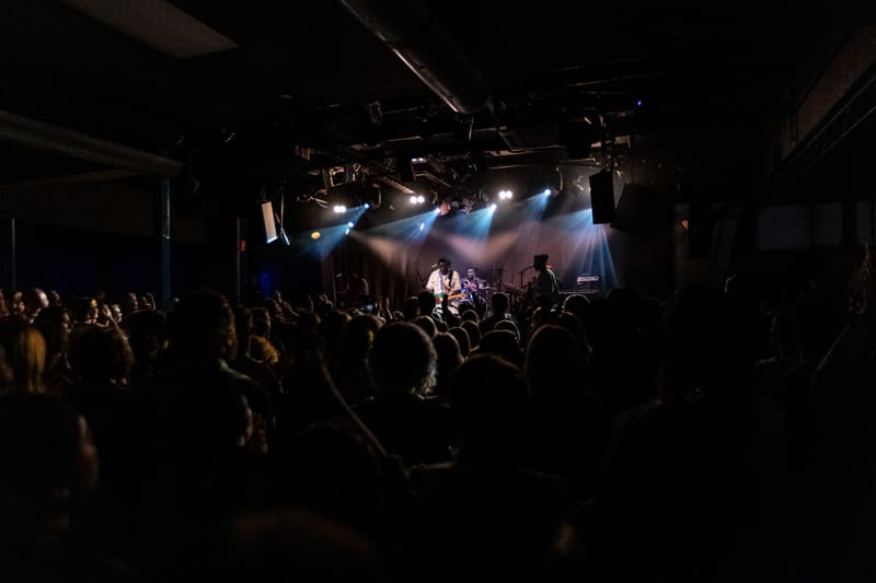Crowd of concert-goers with raised hands silhouetted against a brightly lit stage during a live performance.