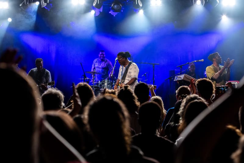 Band performing on stage under blue lighting with audience members' heads visible in foreground.