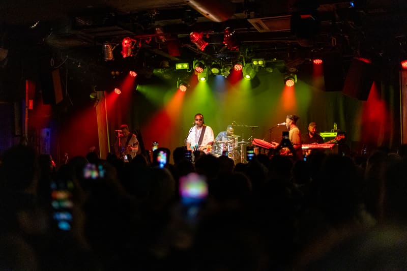 Band performing on stage bathed in red and green spotlight, audience members holding up phones to record.