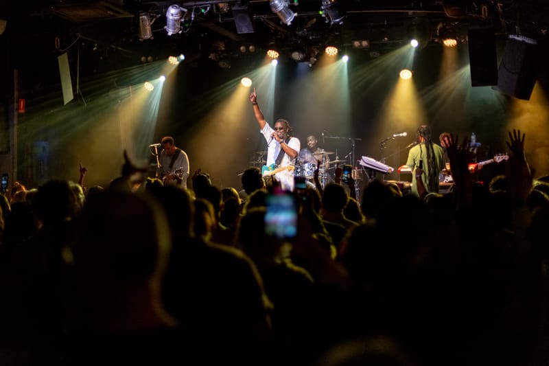 The Original Wailers perform on stage bathed in golden spotlights whilst audience members raise their hands and record the concert.