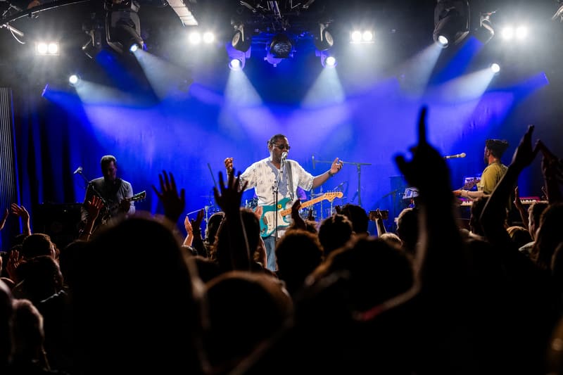 Musician in light blue shirt centre stage with arms raised, surrounded by band members and crowd silhouettes beneath blue stage lighting.
