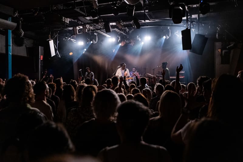 Dense crowd with raised hands facing a stage lit by bright overhead spotlights during a live music performance.