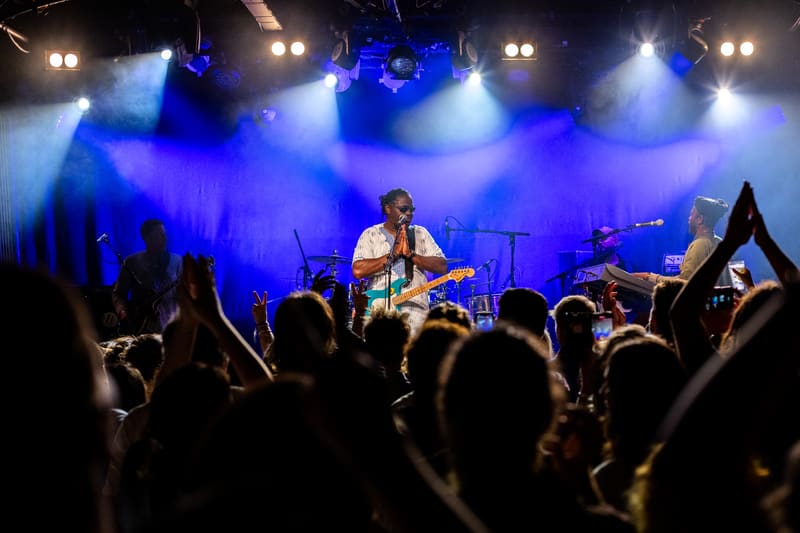 Musician with guitar performs on stage beneath blue spotlights before a crowd of raised hands and phone cameras.