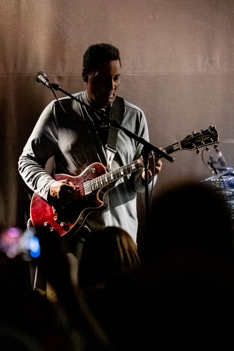 Musician performing with red electric guitar and microphone under stage lighting to an audience.