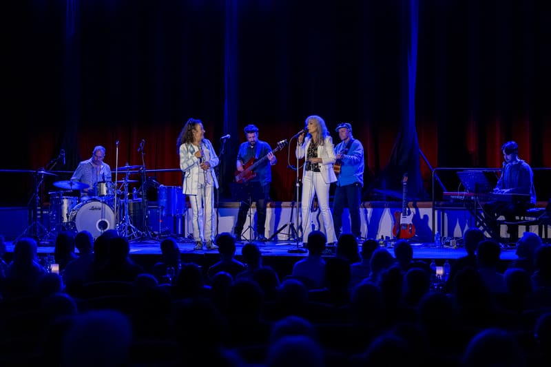 Two female vocalists perform with a live band on stage under blue theatrical lighting, with audience members visible in the foreground.