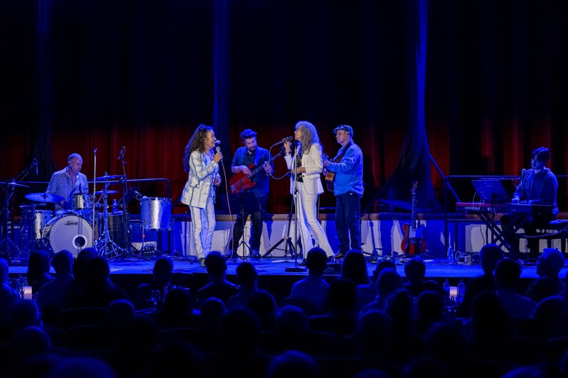 Two female vocalists perform centre stage with live band beneath blue lighting, audience members holding phones in foreground.