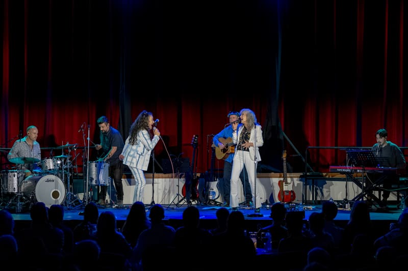 Two female singers perform centre stage with a live band, bathed in blue spotlights against red curtains.
