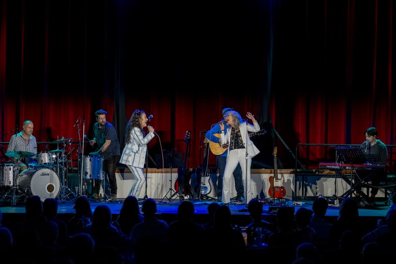 Six musicians perform on a stage lit by red and blue lights, with audience members visible in silhouette below.