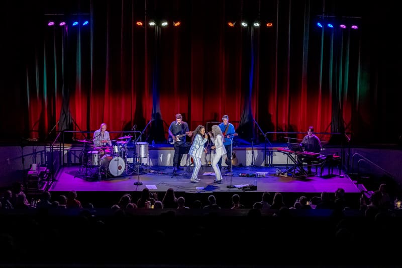 Two female singers perform centre stage with a live band under dramatic red and blue lighting during a concert production.