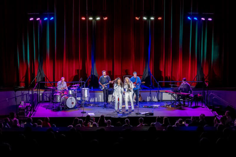 Two female singers perform centre stage with a live band, bathed in purple and red lighting beneath vertical light installations.