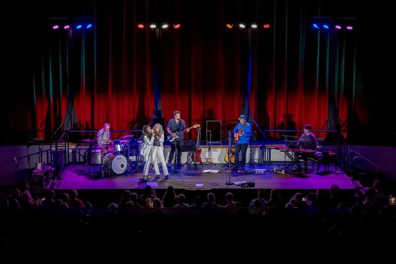 Two singers perform centre stage with a live band under red and blue lighting, audience silhouetted in foreground.