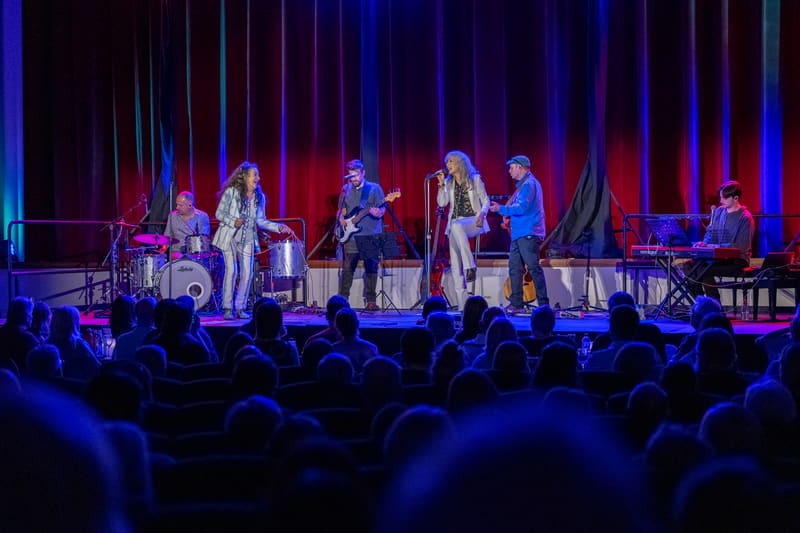 Two female singers and band members perform on a lit stage before an audience in a darkened theatre.