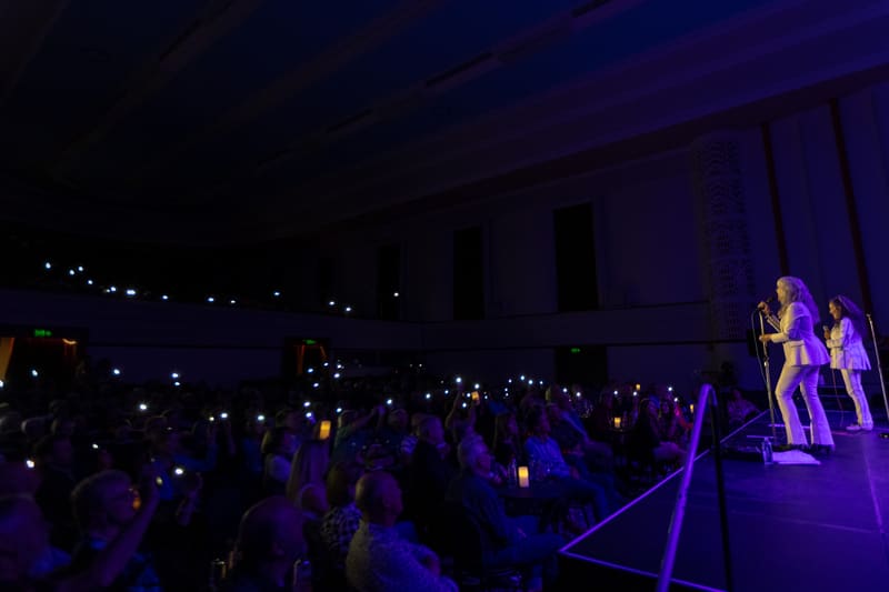 Two performers on a dimly lit stage bathed in purple light, with an engaged audience silhouetted in the foreground.