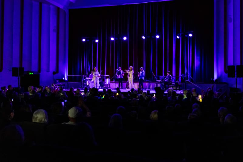 Two performers on stage surrounded by band members, bathed in blue stage lighting with audience silhouettes in foreground.