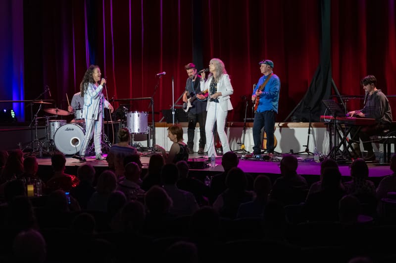Two female performers and band members on stage under purple and blue lighting during the We're Going To Graceland concert.