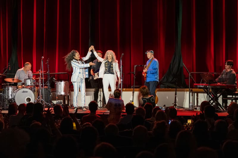 Two performers in white outfits raise their hands together centre stage beneath red curtains, with band members and audience silhouettes visible.