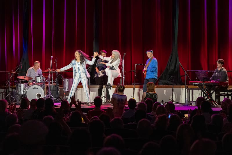 Two female performers in white outfits centre stage, surrounded by band members, with audience silhouettes in foreground under purple stage lighting.