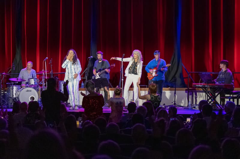 Two female vocalists in flowing white outfits perform centre stage with a full band beneath red theatrical lighting, audience silhouettes in foreground.