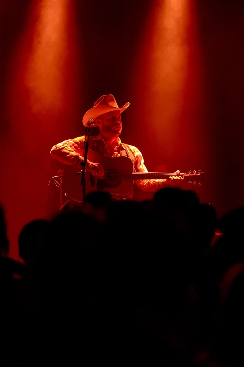 Guitarist in a cowboy hat performing on stage beneath warm amber spotlights, audience silhouettes in foreground.