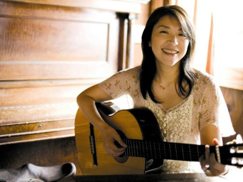 Woman with dark hair holding an acoustic guitar, smiling in warm indoor light.