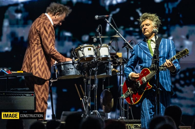 Two musicians perform on stage, one at keyboards in a brown patterned jacket and one with a guitar in a blue striped jacket, during Split Enz tour.