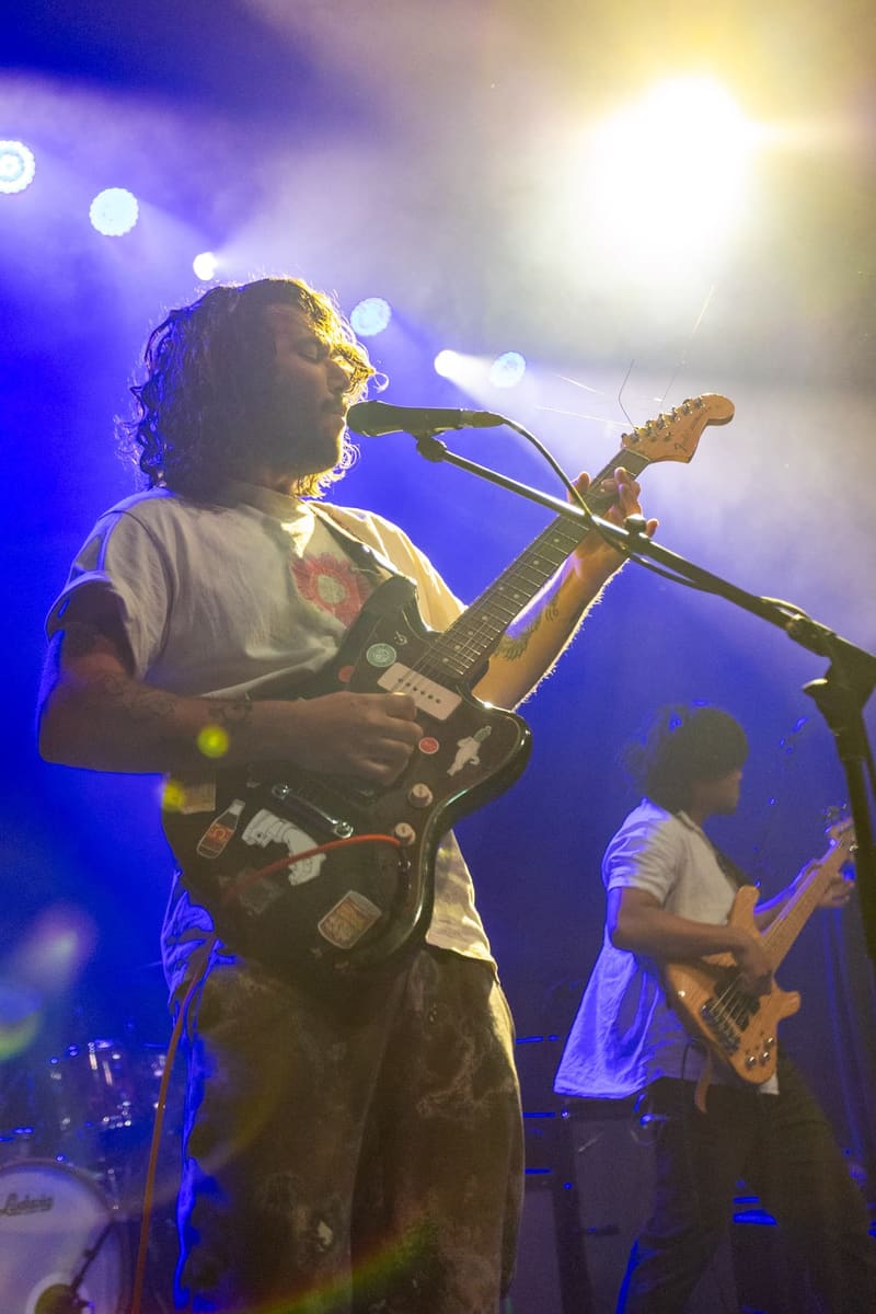 Guitarist singing into microphone under blue and white stage lighting, with bassist visible behind.