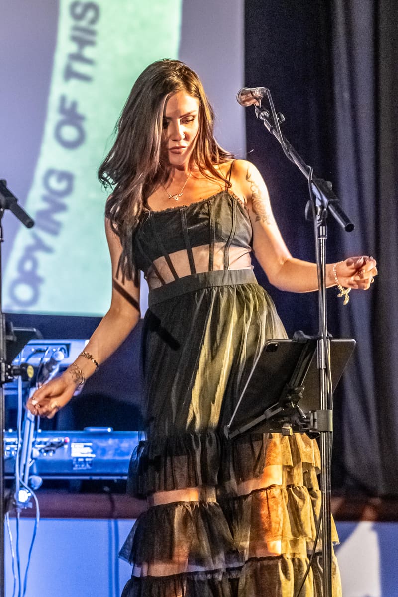 Female performer in a black and gold layered dress singing at a microphone during a stage performance.
