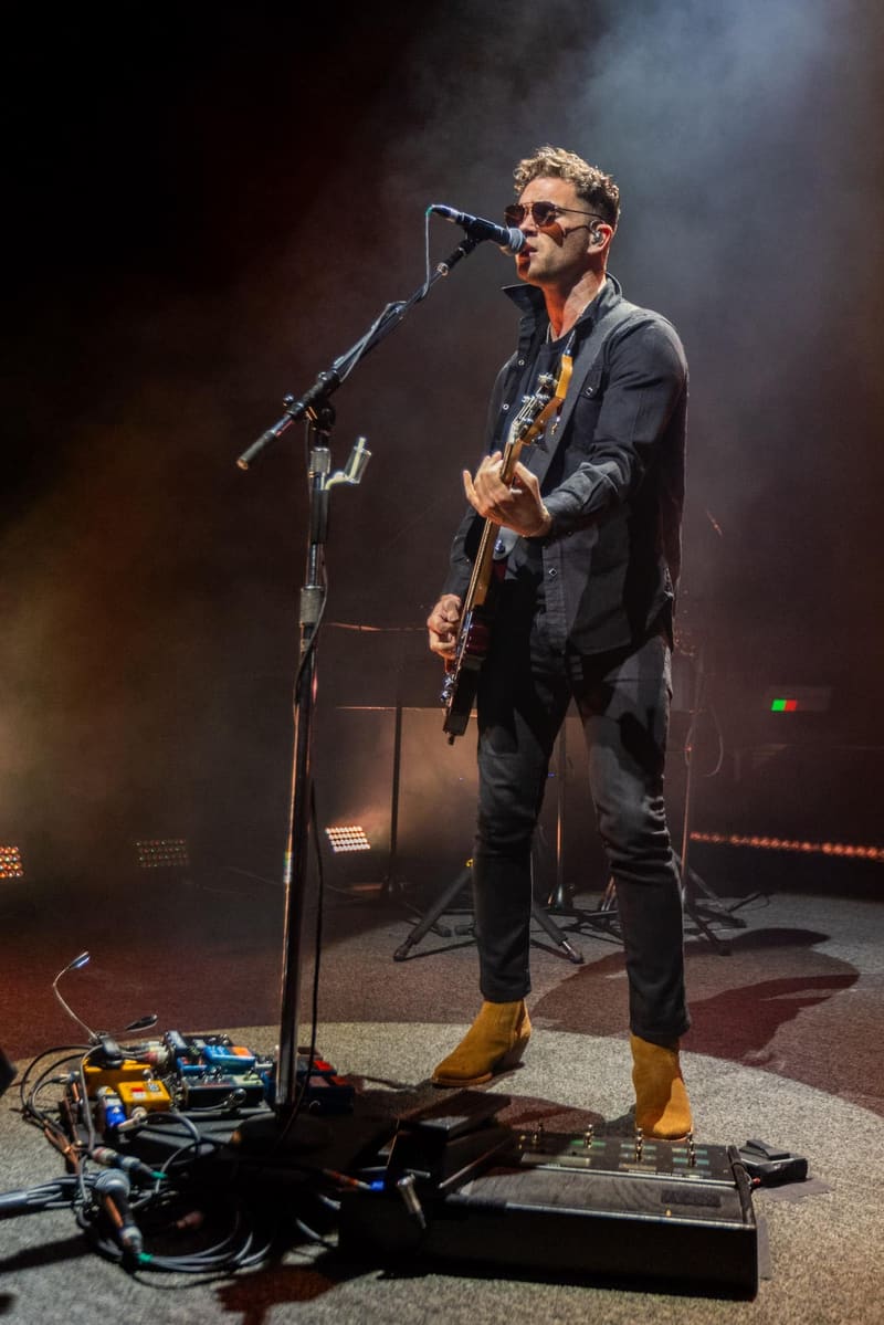 Guitarist and vocalist performing at a microphone on stage with amplification equipment and warm stage lighting.