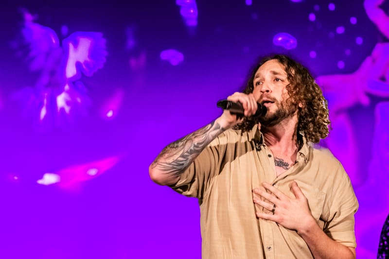 Man with curly hair singing into microphone against vibrant purple stage lighting and bokeh background.