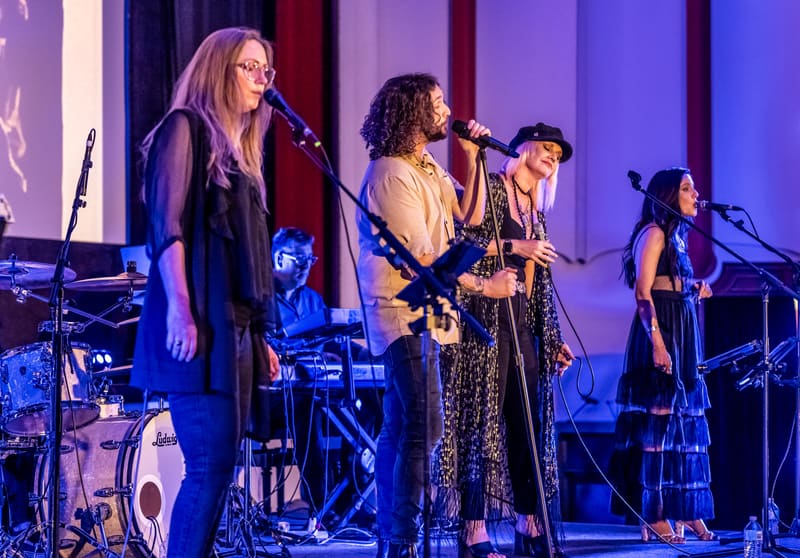 Four musicians perform on stage under purple lighting, with vocals, guitar, and drums visible in an ensemble arrangement.