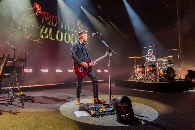 Guitarist in dark jacket performing on illuminated stage with "Royal Blood" backdrop and drummer visible behind.