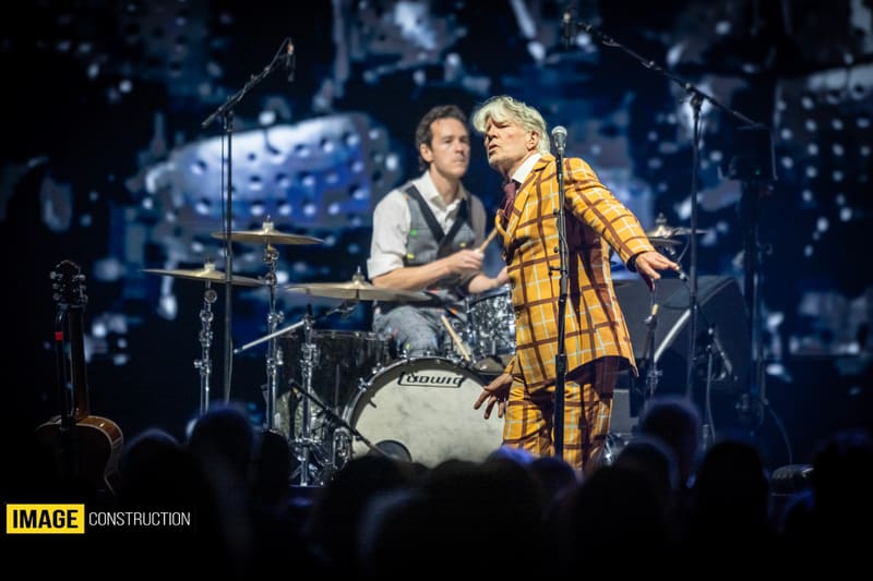 Two performers on stage during Split Enz concert, one in yellow jacket singing, one playing guitar, with drum kit and blue stage lighting.