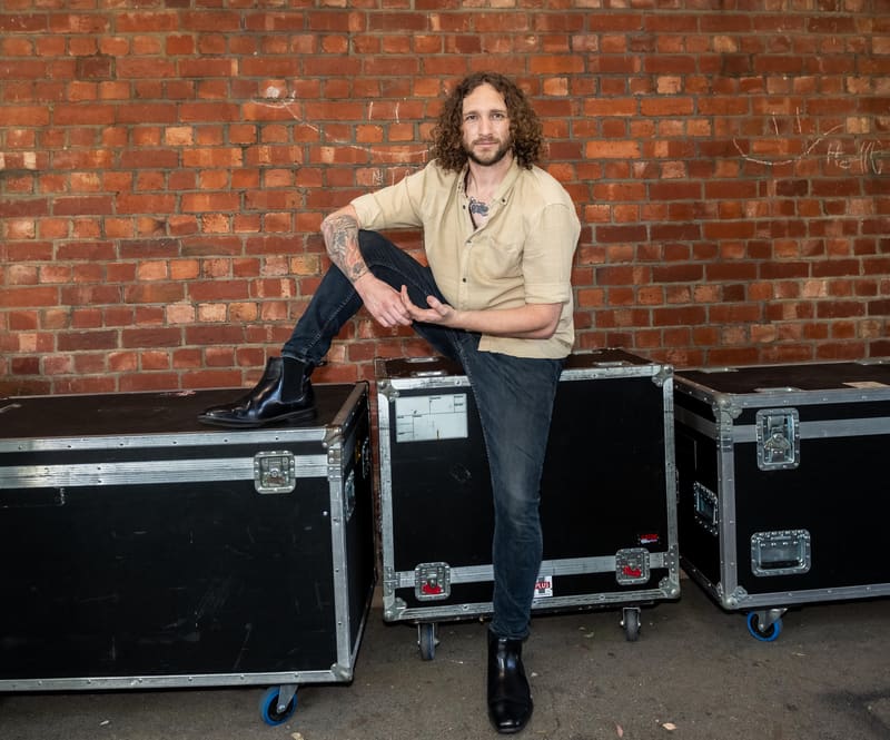 Man with long curly hair seated on black flight cases against a red brick wall.