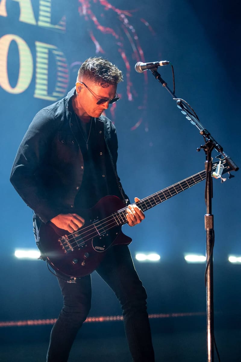 Bassist in dark clothing performing at a microphone stand under blue stage lighting during a Royal Blood concert.