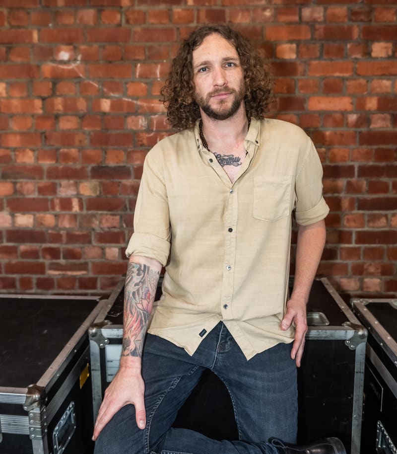 Man with curly hair and tattooed arm seated against exposed red brick wall in gallery setting.