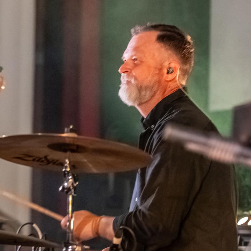 Drummer in black shirt performing at drums with cymbal in motion during gallery event.